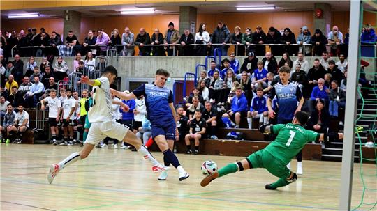 Hallenszene eines Fußballspiels mit mehreren Spielern in Aktion, ein Spieler in grün rutscht zum Ball, Zuschauer sitzen auf Tribünen im Hintergrund