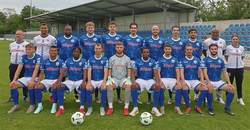 Mannschaftsfoto einer Fußballmannschaft mit Spielern in blauen Trikots und Personen in weißen Trainingsanzügen auf einem Fußballfeld vor einer Tribüne