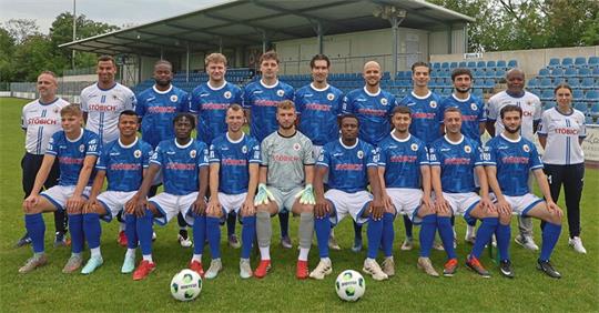 Mannschaftsfoto einer Fußballmannschaft mit Spielern in blauen Trikots und Personen in weißen Trainingsanzügen auf einem Fußballfeld vor einer Tribüne