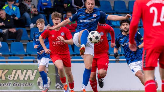 Fußballspieler in blauen und roten Trikots kämpfen um den Ball auf dem Spielfeld vor Zuschauern auf der Tribüne