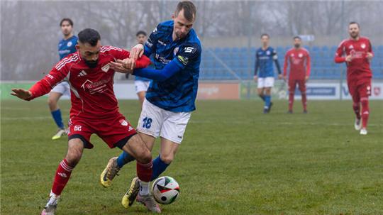 Zwei Fußballspieler kämpfen um den Ball, einer in rotem Trikot, der andere in blauem Trikot mit weißer Hose, auf einem Spielfeld mit weiteren Spielern im Hintergrund.