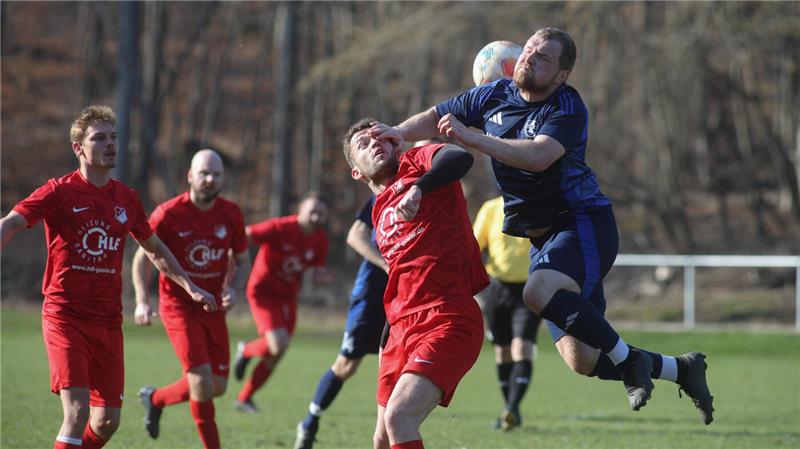 Fußballspieler in roten und blauen Trikots springen und kämpfen um den Ball in der Luft auf einem Spielfeld mit unscharfem Waldhintergrund