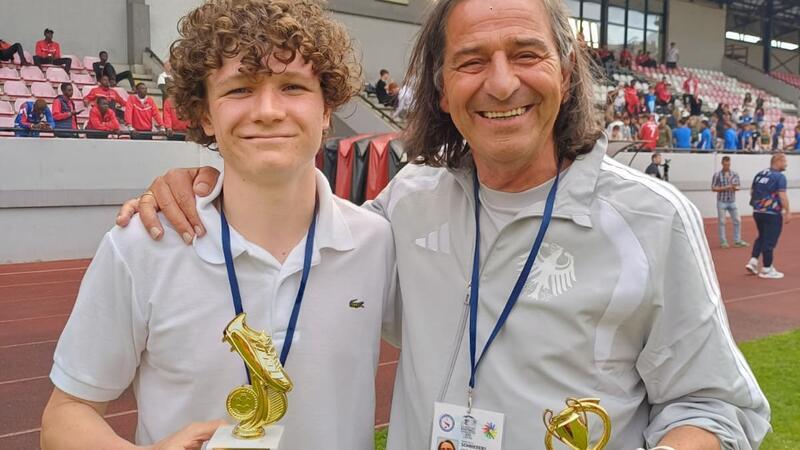 Zwei Personen mit Medaillen um den Hals halten jeweils eine goldfarbene Trophäe auf einem Sportplatz, im Hintergrund Tribünen mit Zuschauern.