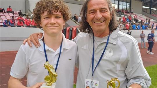 Zwei Personen mit Medaillen um den Hals halten jeweils eine goldfarbene Trophäe auf einem Sportplatz, im Hintergrund Tribünen mit Zuschauern.