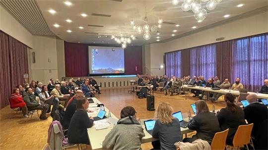 Großer Sitzungssaal mit mehreren Personen, die an Tischen mit Laptops sitzen und einer Präsentation auf einer Leinwand folgen
