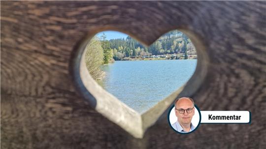 Blick durch ein herzförmiges Loch in einem Holzbrett auf einen See mit bewaldetem Ufer