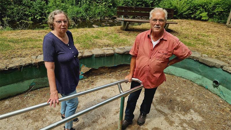 Eine Frau und ein Mann stehen an einem leeren, runden Becken mit grün gestrichener Innenwand und Metallgeländer im Freien, das stark sanierungsbedürftig ist.