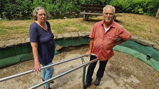 Eine Frau und ein Mann stehen an einem leeren, runden Becken mit grün gestrichener Innenwand und Metallgeländer im Freien, das stark sanierungsbedürftig ist.