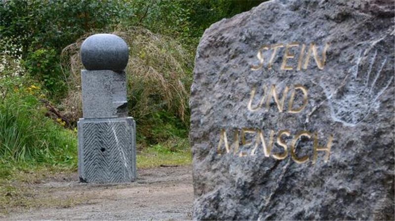 Großer Fels mit der goldfarbenen Inschrift 'STEIN UND MENSCH' neben einer abstrakten Steinskulptur in einer grünen Parklandschaft