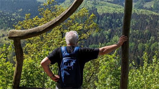 Rückansicht eines Mannes mit grauen Haaren und blauem Rucksack, der mit einer Hand an einem Holzpfosten lehnt und auf bewaldete Hügel blickt.