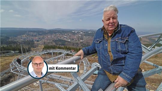 Person mit Jeansjacke lehnt an einem Metallgeländer mit Blick auf eine kurvige Sommerrodelbahn und eine Stadt im Hintergrund