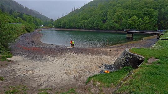Flussufer mit Kiesstrand, zwei Personen in Warnwesten am Wasser und bewaldete Hügel im Hintergrund