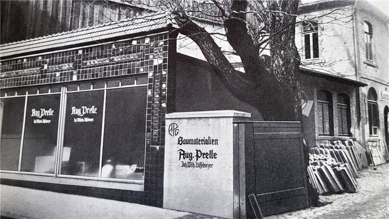 Schwarz-weiß-Fotografie eines Gebäudes mit Schaufenstern und einem Schild mit der Aufschrift 'Aug. Prelle' neben einem großen Baum