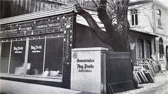 Schwarz-weiß-Fotografie eines Gebäudes mit Schaufenstern und einem Schild mit der Aufschrift 'Aug. Prelle' neben einem großen Baum