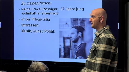 Ein Mann steht vor einem Bildschirm, auf dem er auch zu sehen ist.