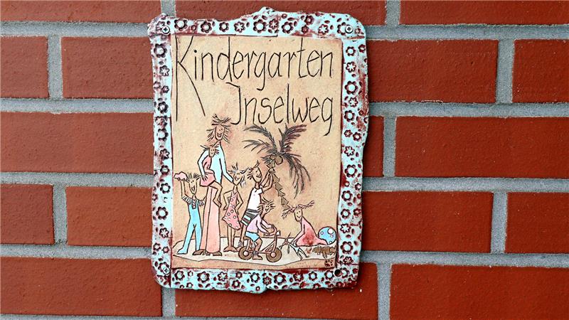 Handgemaltes Schild mit der Aufschrift 'Kindergarten Inselweg' und einer Illustration von Kindern vor einer Palme an einer roten Backsteinwand
