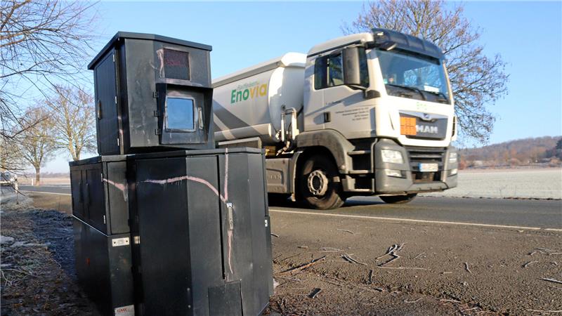 Schwarze Geschwindigkeitsmessanlage am Straßenrand vor einem vorbeifahrenden weißen Tanklastwagen.