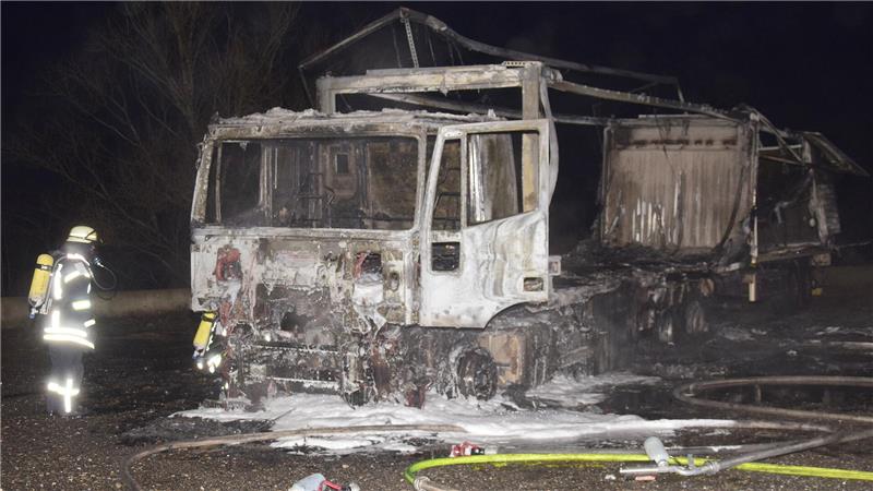 Ausgebrannter Lastwagen auf einer Straße bei Nacht mit Feuerwehrmann in Schutzkleidung daneben