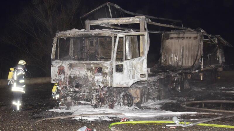 Ausgebrannter Lastwagen auf einer Straße bei Nacht mit Feuerwehrmann in Schutzkleidung daneben
