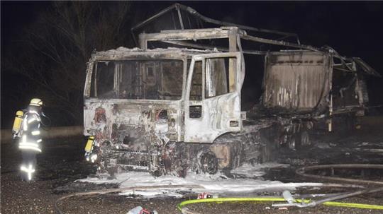 Ausgebrannter Lastwagen auf einer Straße bei Nacht mit Feuerwehrmann in Schutzkleidung daneben