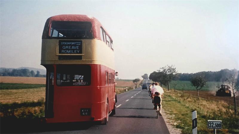 Roter Doppeldeckerbus auf Landstraße mit Radfahrern rechts daneben und Feldern im Hintergrund.