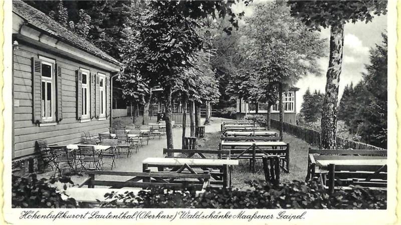 Außenbereich eines Gasthauses mit mehreren Holztischen und Stühlen unter Bäumen neben einem langgestreckten Gebäude.