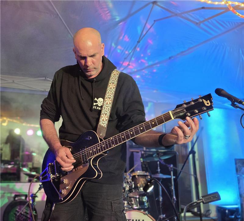 Person in schwarzem Kapuzenpullover spielt E-Gitarre auf einer Bühne mit Schlagzeug im Hintergrund