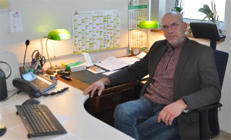 Mann in Sakko und Jeans sitzt an einem Schreibtisch mit Computer, Kalendern und zwei grünen Schreibtischlampen in einem Büro mit weißen Wänden und Fenster mit Orchidee