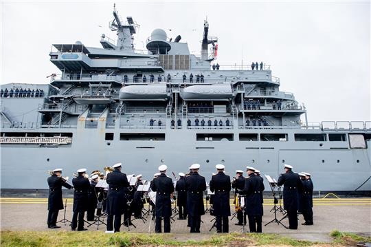 Der Marinemusikkorps Wilhelmshaven steht an einer Kaimauer, während die „Frankfurt am Main“ in den Hafen einfährt.