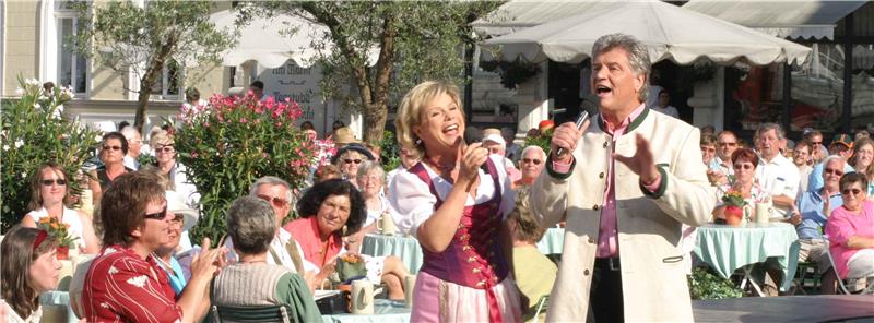 Zwei Personen singen mit Mikrofonen vor einer sitzenden Menschenmenge im Freien unter Sonnenschirmen