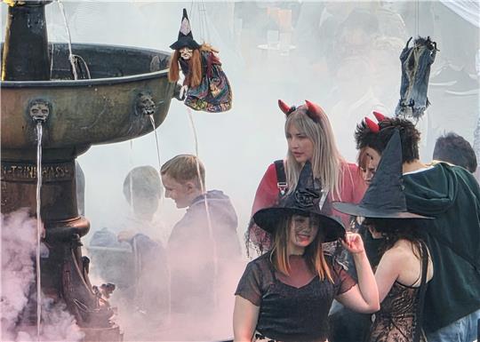 Gruppe von Menschen in Halloween-Kostümen mit Hexenhut und Teufelshörnern vor einem Brunnen mit Dampf und einer Hexenpuppe. 
