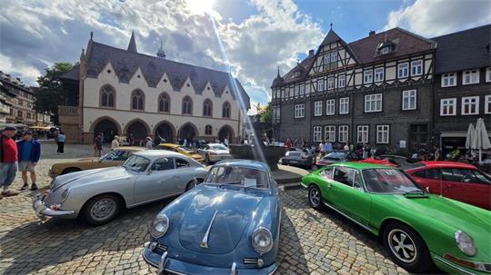 Mehrere klassische Sportwagen auf einem gepflasterten Platz vor historischen Fachwerkhäusern und einer Kirche bei sonnigem Himmel