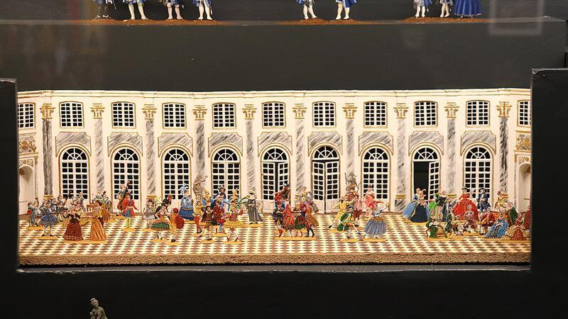 Miniaturmodell eines prunkvollen Saals mit zahlreichen Figuren in historischen Kostümen auf einem Schachbrettmusterboden vor einer Wand mit großen Fenstern und Verzierungen