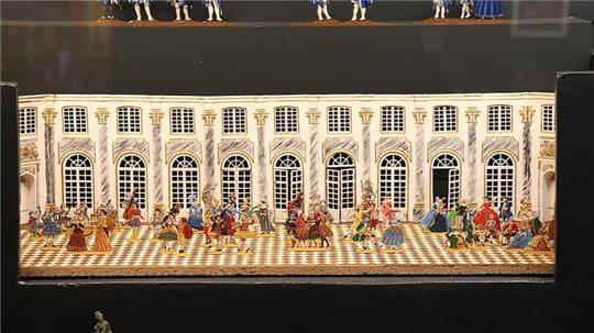 Miniaturmodell eines prunkvollen Saals mit zahlreichen Figuren in historischen Kostümen auf einem Schachbrettmusterboden vor einer Wand mit großen Fenstern und Verzierungen