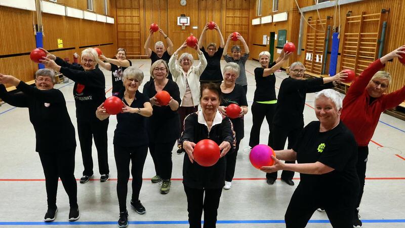 Gruppe älterer Menschen in Sportkleidung steht in einer Turnhalle und hält rote Gymnastikbälle vor sich oder über dem Kopf.