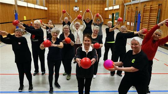 Gruppe älterer Menschen in Sportkleidung steht in einer Turnhalle und hält rote Gymnastikbälle vor sich oder über dem Kopf.