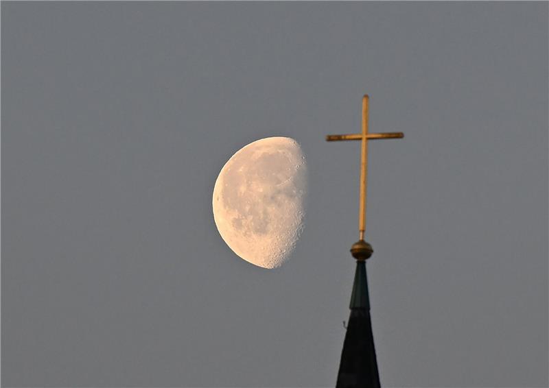 Kirchenbindung und die Religiosität gehen zurück Der Mond im Morgenlicht hinter einem Kreuz auf einem Kirchturm.