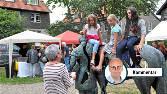 Drei Mädchen sitzen auf einer bronzenen Skulptur eines Pferdes, davor steht eine ältere Frau mit kurzen grauen Haaren und gestreiftem Oberteil, im Hintergrund Zelte und weitere Personen auf einem Marktplatz.