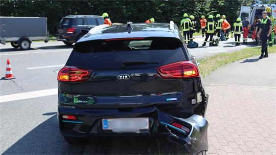Ein am Heck beschädigter Kia steht am Straßenrand. Im Hintergrund sind ein Krankenwagen und Feuerwehrleute zu sehen.