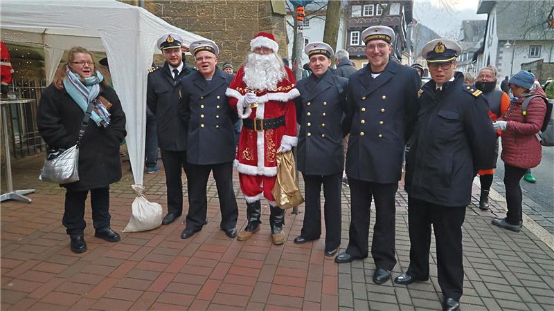 Am Super-Samstag kommen Besucher aus ganz Deutschland Mehrere Männer in Marineuniformen stehen neben einer Person in einem roten Weihnachtsmannkostüm mit goldenem Sack auf einem gepflasterten Platz mit Marktständen im Hintergrund.