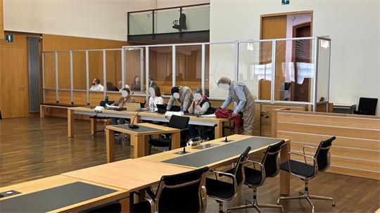 Innenraum eines Sitzungssaals mit mehreren Personen hinter Plexiglasscheiben an einem langen Tisch und weiteren leeren Tischen mit Stühlen im Vordergrund.