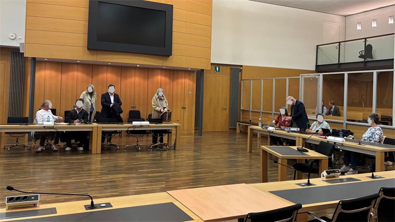 Mehrere Personen sitzen mit Abstand in einem großen Raum mit Holzboden und Tischen, einige tragen Mund-Nasen-Schutz.