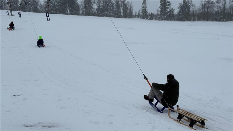 Das Foto zeigt einen Mann auf einem Schlitten, der sich von einem Lift den Hang hinauziehen lässt und einen weiteren Schlitten hinter sich her zieht.