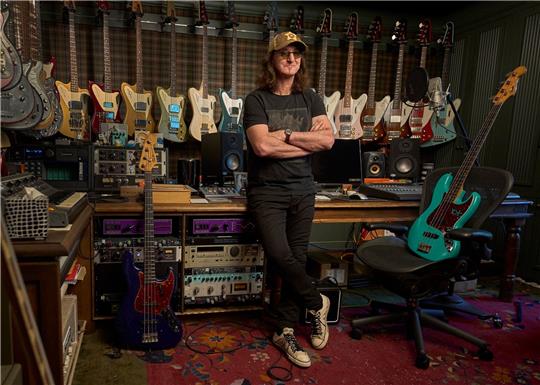 Der Rush-Frontmann Geddy Lee steht in seinem Studio, Bassgitarren hängen an der Wand.