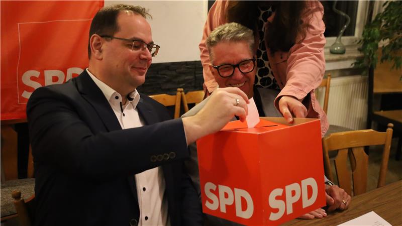 Person in dunklem Sakko wirft Zettel in rote Wahlurne mit SPD-Logo, daneben steht eine weitere Person in rosa Jacke