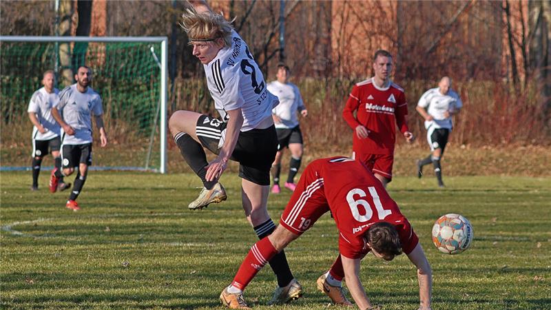 Zwei Fußballspieler in Aktion auf dem Spielfeld, einer in weißem Trikot springt, der andere in rotem Trikot beugt sich nach vorne, im Hintergrund weitere Spieler und ein Tor
