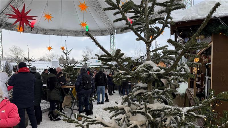 Hinter einer schneebedeckten Fichte stehen viele Menschen an Bistrotischen und Ständen.