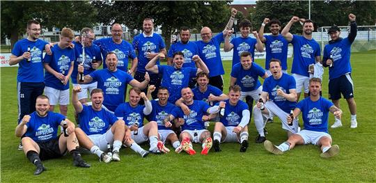 Gruppe von Fußballspielern in blauen Trikots jubelnd auf dem Spielfeld vor Bäumen und bewölktem Himmel