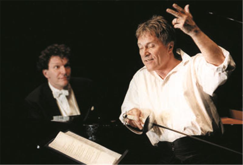 Ein Schauspieler mit Degen und weißem Hemd hebt den Arm zu einer Pose; im Hintergrund blickt ihn eine Klavierspieler mit großen Augen an.