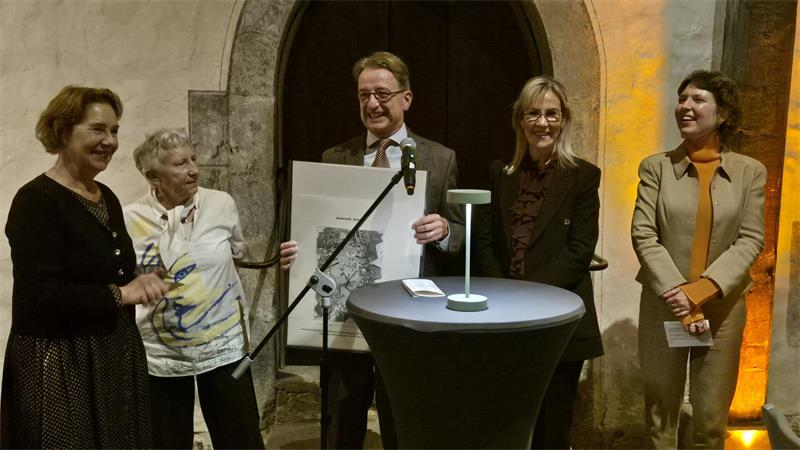 Um einen Stehtisch sind fünf Menschen versammelt, vier Frauen und ein Mann, der Mann hält ein Plakat in Händen, alle lächeln.